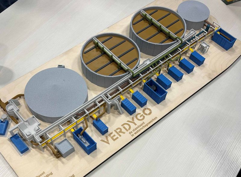 Model van een waterzuiveringsinstallatie met drie grote ronde tanks en een reeks blauwe en gele technische componenten verbonden door buizen en leidingen op een houten plaat met het logo 'VERDYGO de standaard in waterzuiveringswerken'.
