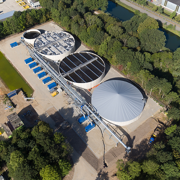 Luchtfoto van een waterzuiveringsinstallatie in een groen gebied met grote ronde tanks en technische apparatuur.