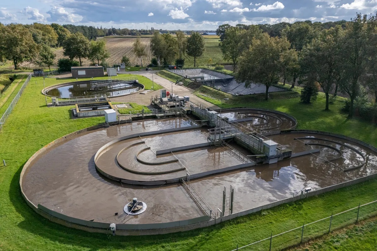Luchtfoto van een waterzuiveringsinstallatie met meerdere ronde bassins gevuld met troebel water, omgeven door grasveld en bomen, onder een partly bewolkte lucht.
