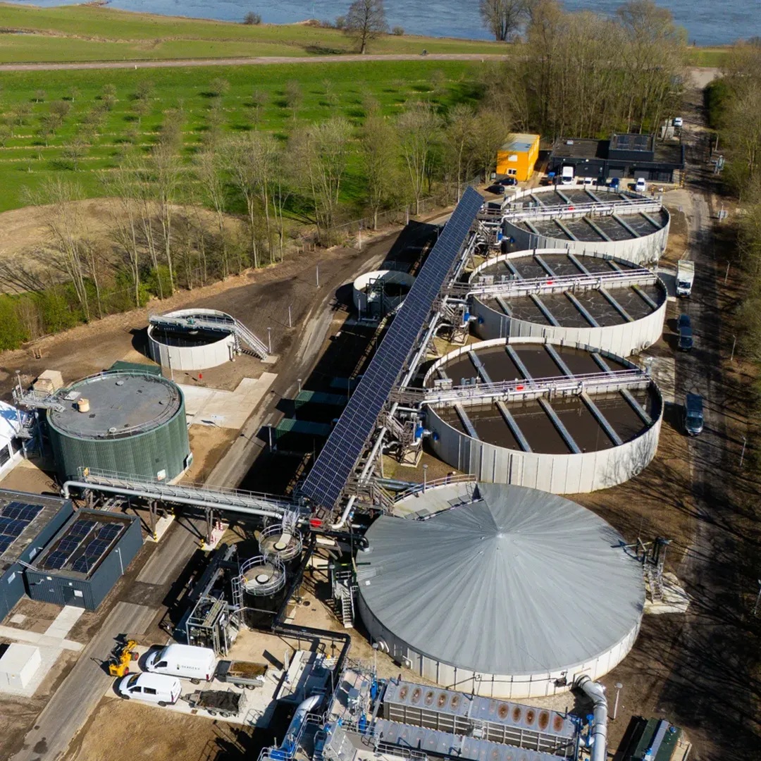 Luchtfoto van een waterzuiveringsinstallatie met meerdere grote, ronde tanks en een rij zonnepanelen. Er zijn wegen en voertuigen rondom, met grasvelden en bomen op de achtergrond langs een rivier.