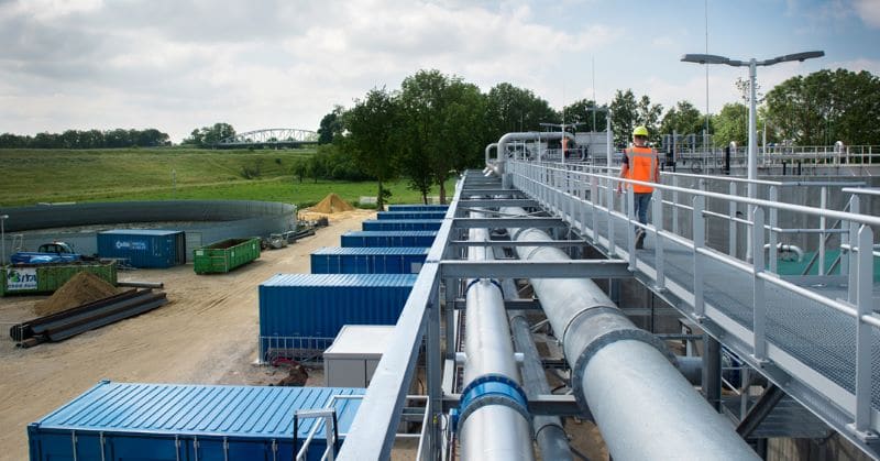 Werkers met veiligheidshelmen en oranje hesjes lopen op een verhoogde metalen catwalk boven grote industriële pijpleidingen in een waterbehandelings- of industriële faciliteit, met blauwe opslagcontainers en een groene omgeving op de achtergrond.