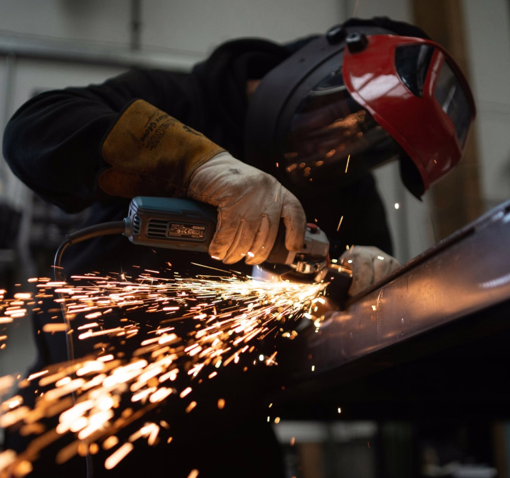 Person wearing protective gloves and face shield using grinder, sparks flying from metal workpiece.