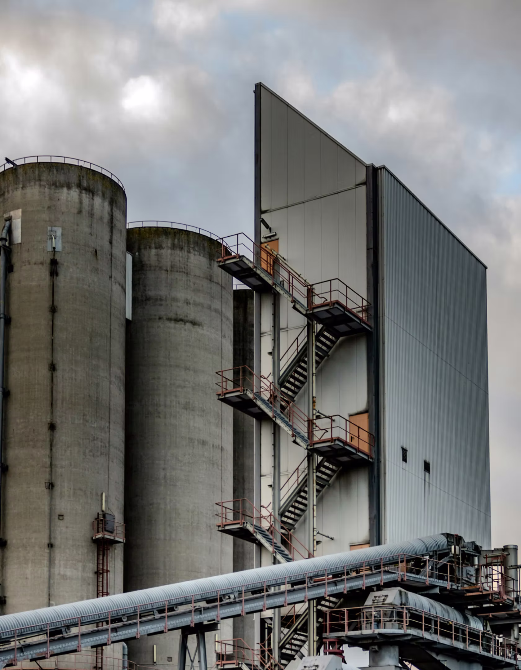 Industrial concrete silos with external staircases and metal conveyor structures under a cloudy sky.