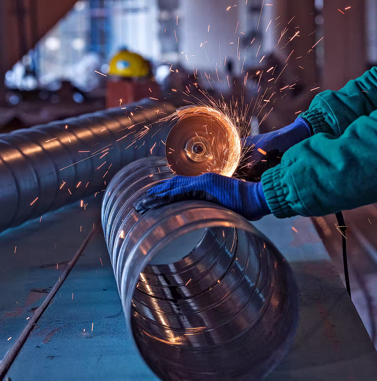 Worker in blue gloves grinding the edge of a metal pipe with sparks flying in a factory setting.