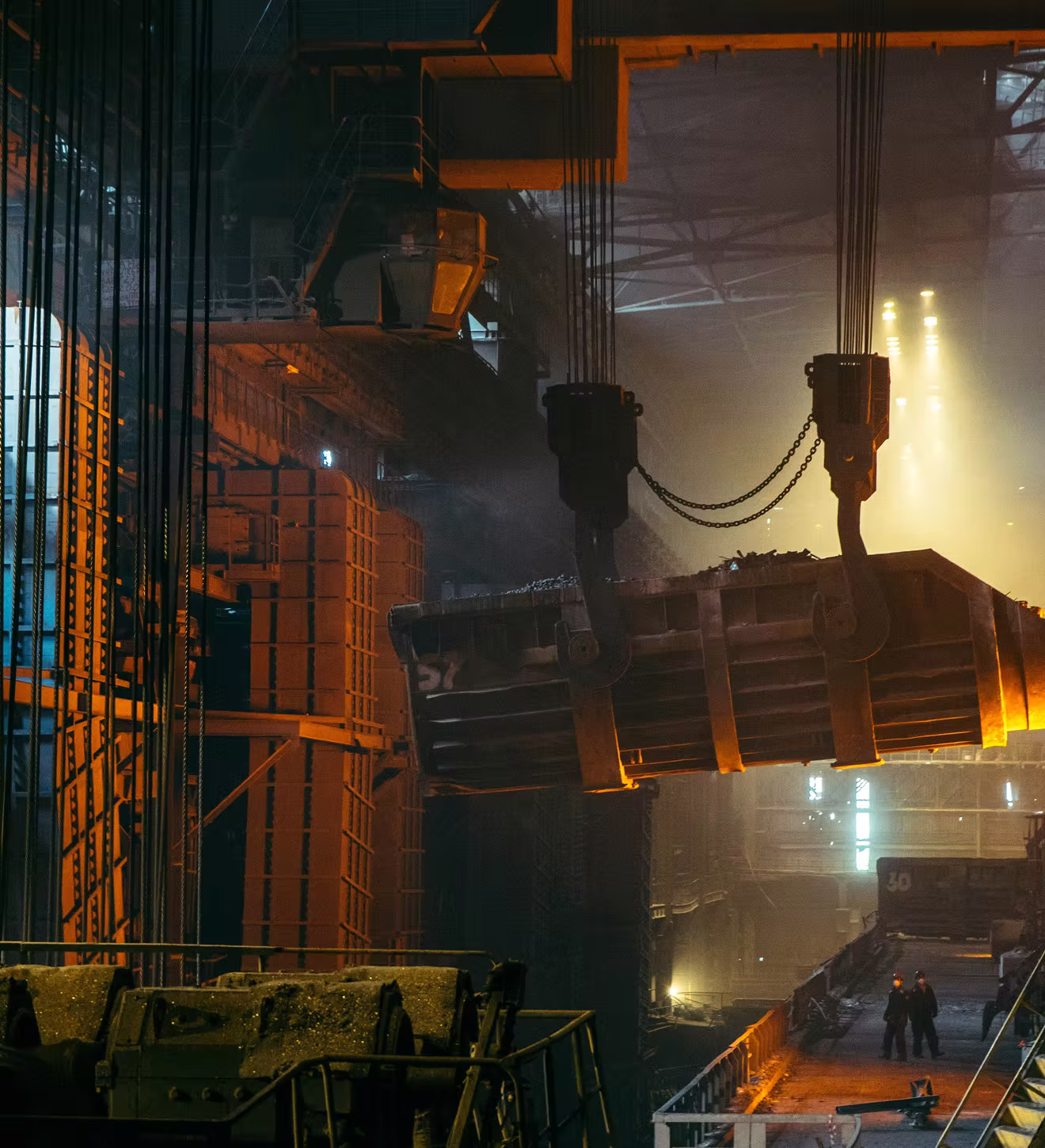 Large industrial crane lifting a heavy load inside a dimly lit factory with two workers below.