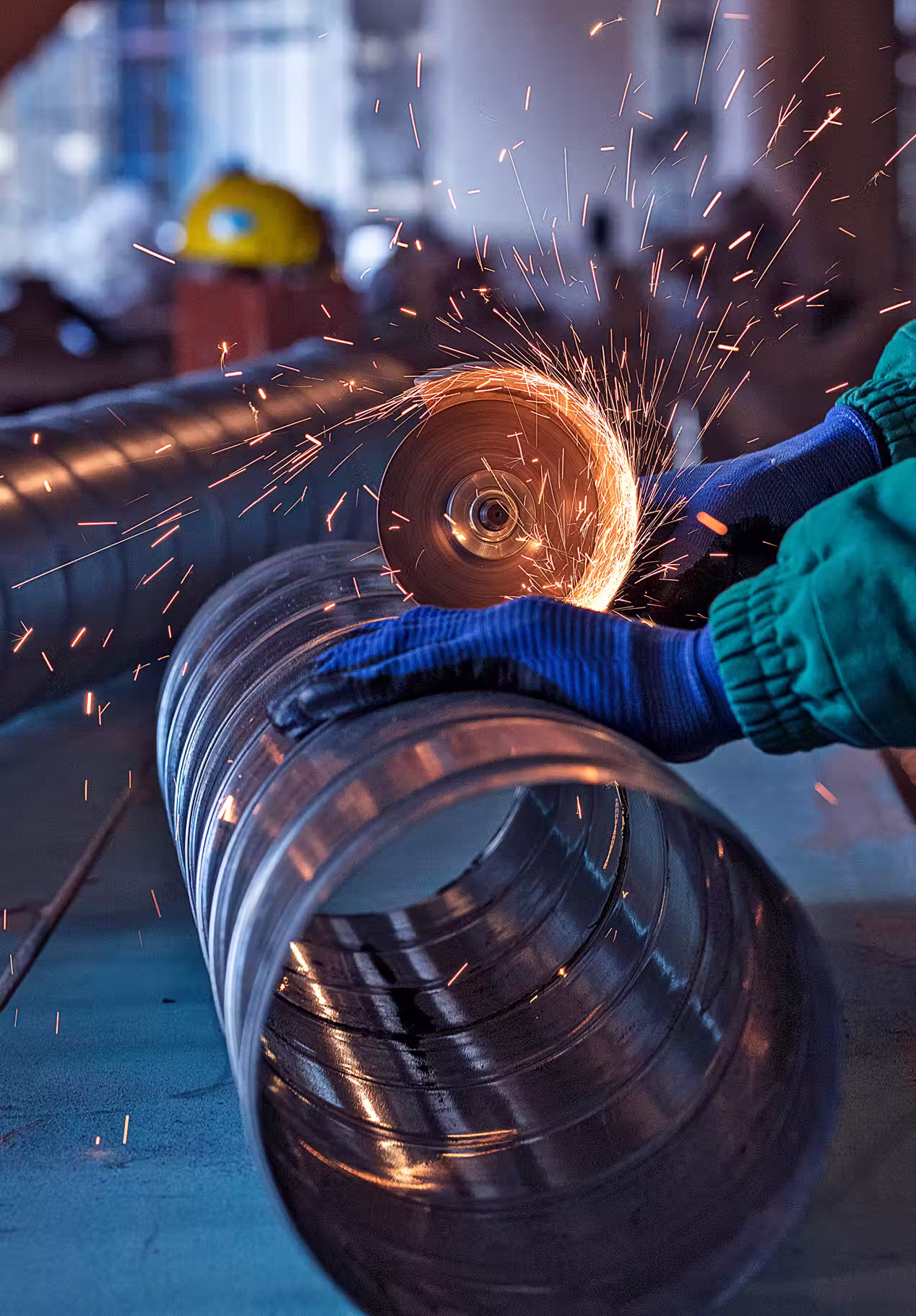 Worker wearing gloves grinding metal pipe with sparks flying in a factory setting.