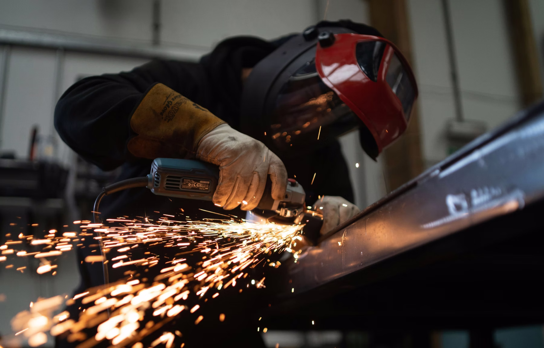 Worker wearing protective gloves and face shield grinding metal with an angle grinder, sparks flying.