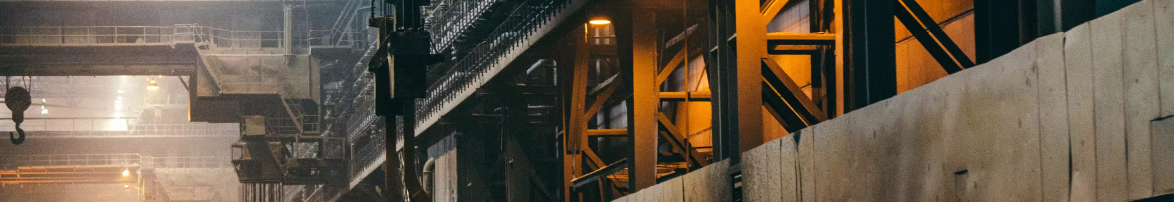 Industrial factory interior with metal beams, stairs, crane hook, and yellow lighting.
