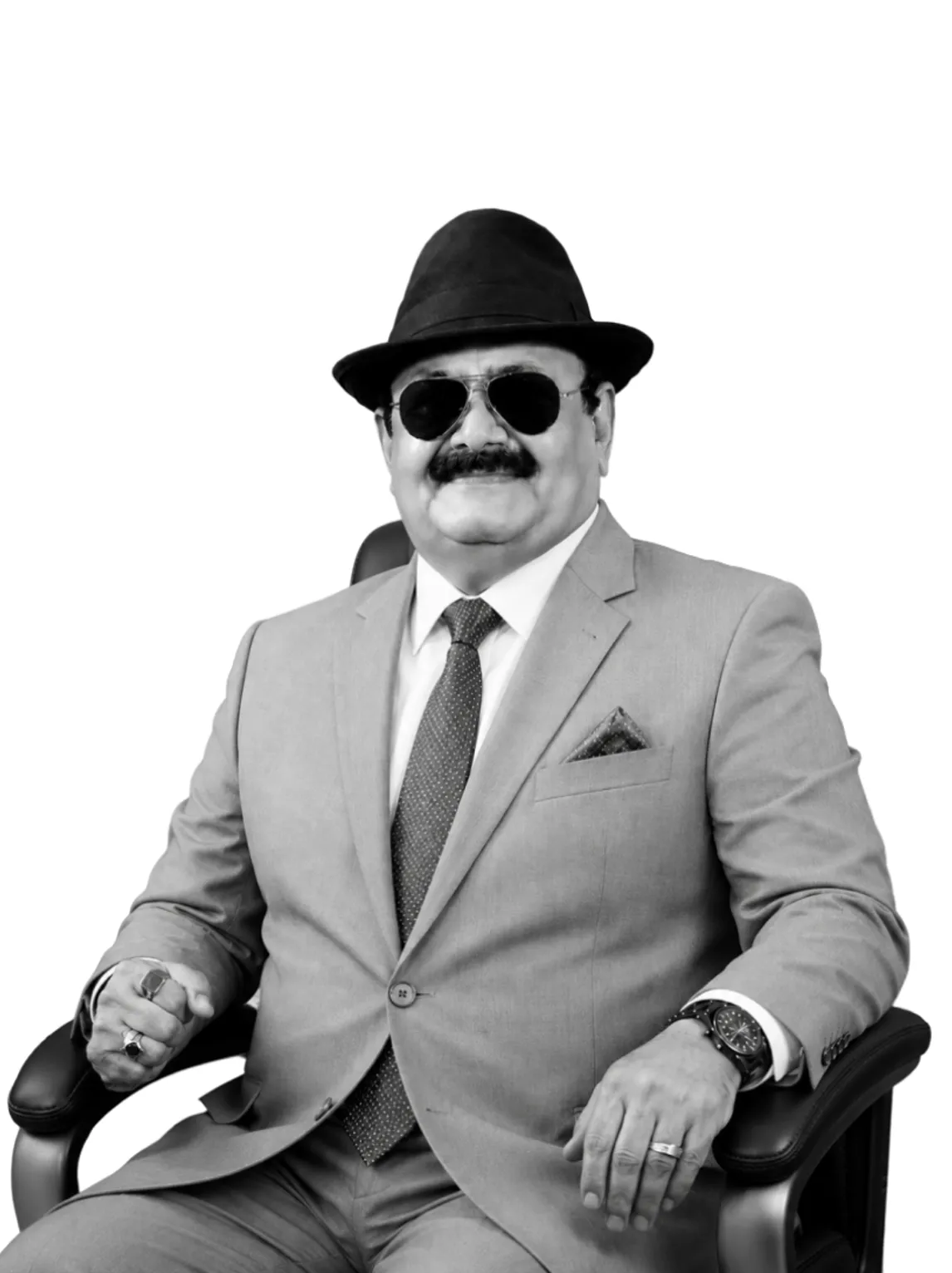 Man in a light suit, tie, sunglasses, and hat seated in a chair, smiling with rings and a watch.