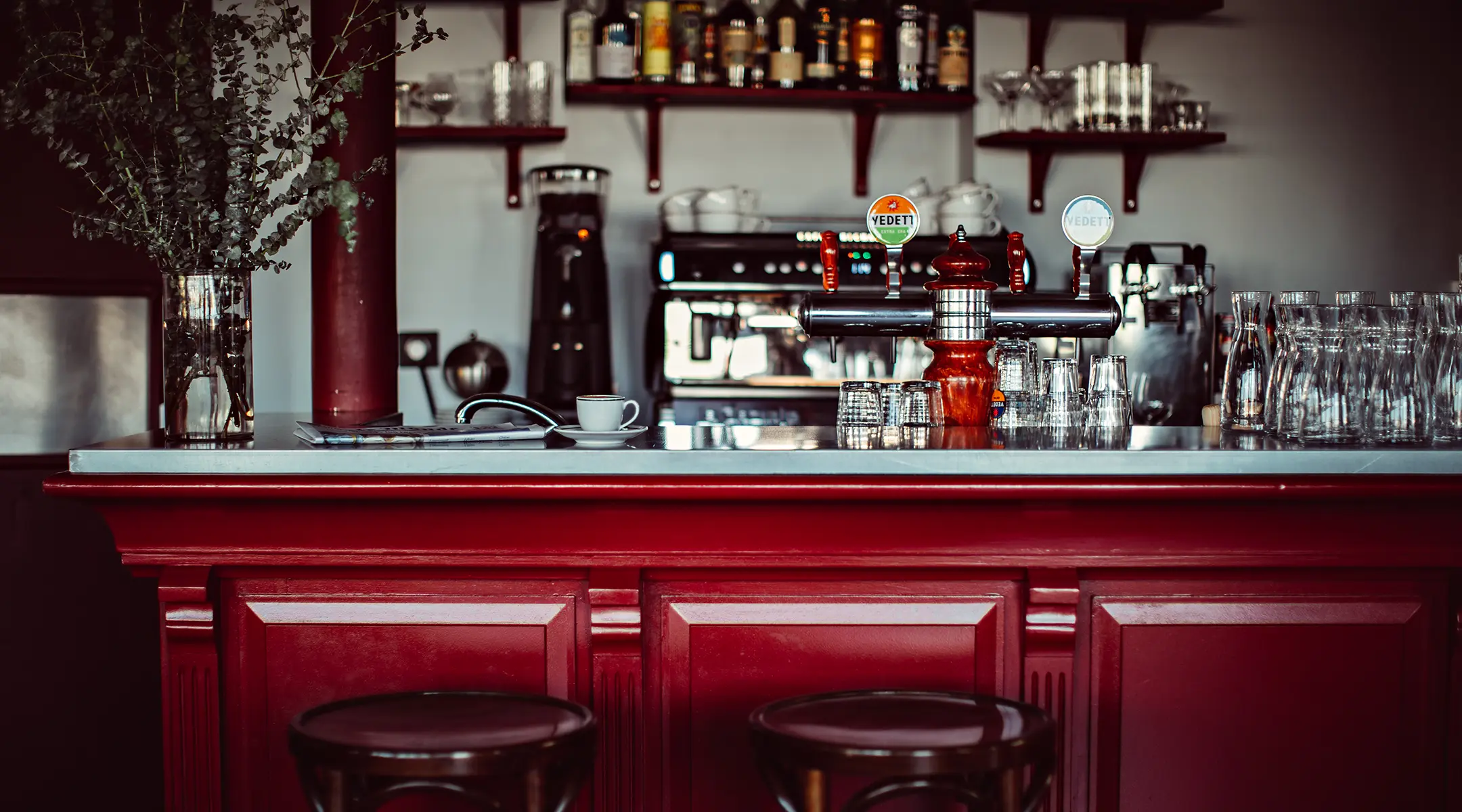 Comptoir de bar bistrot en bois rouge et zinc avec tireuse à bière, machine à expresso, tasse de café et étagères garnies de bouteilles et de verres, devant deux tabourets