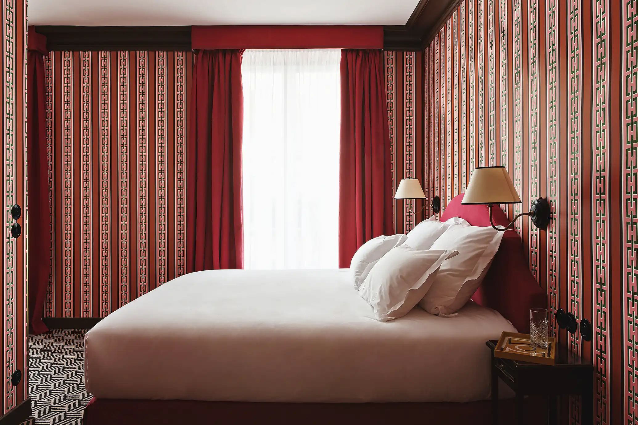 Cozy room featuring red and orange geometric-striped wallpaper, a double bed shown in profile, red velvet curtains, and a window with sheer white drapes