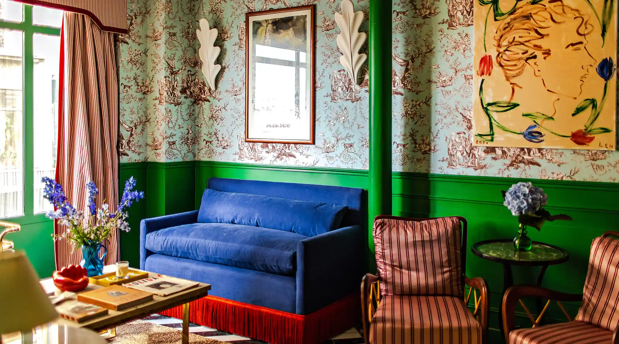 Living room corner featuring a bouquet of blue delphiniums on a coffee table stacked with books, set beside a blue velvet sofa with red fringe and striped curtains.