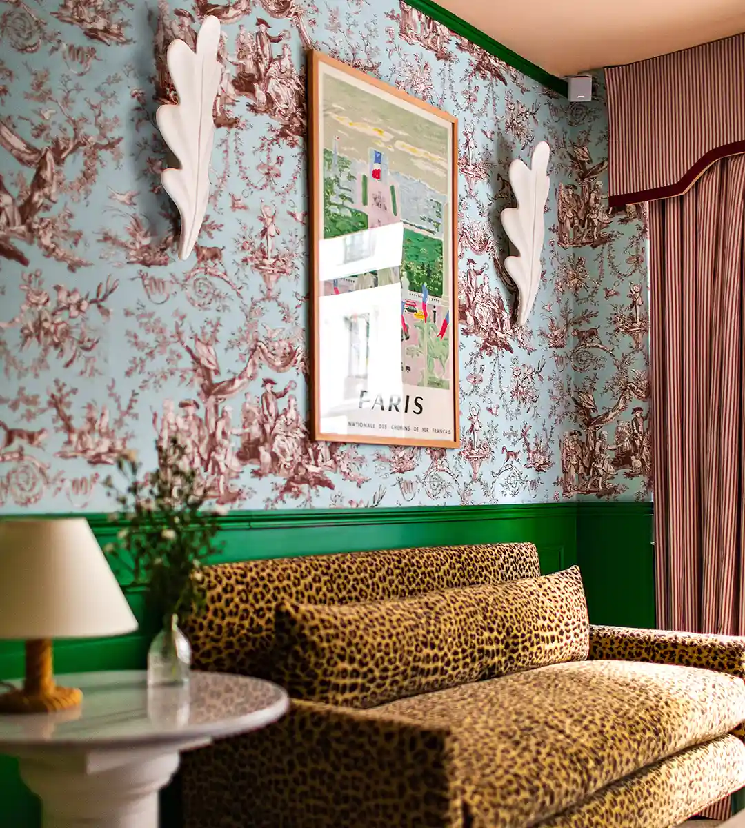 Canapé imprimé léopard devant un mur en toile de Jouy bleu et bordeaux, encadré d'appliques en forme de feuille et d'une affiche vintage des chemins de fer sur Paris, avec lampe sur guéridon en marbre