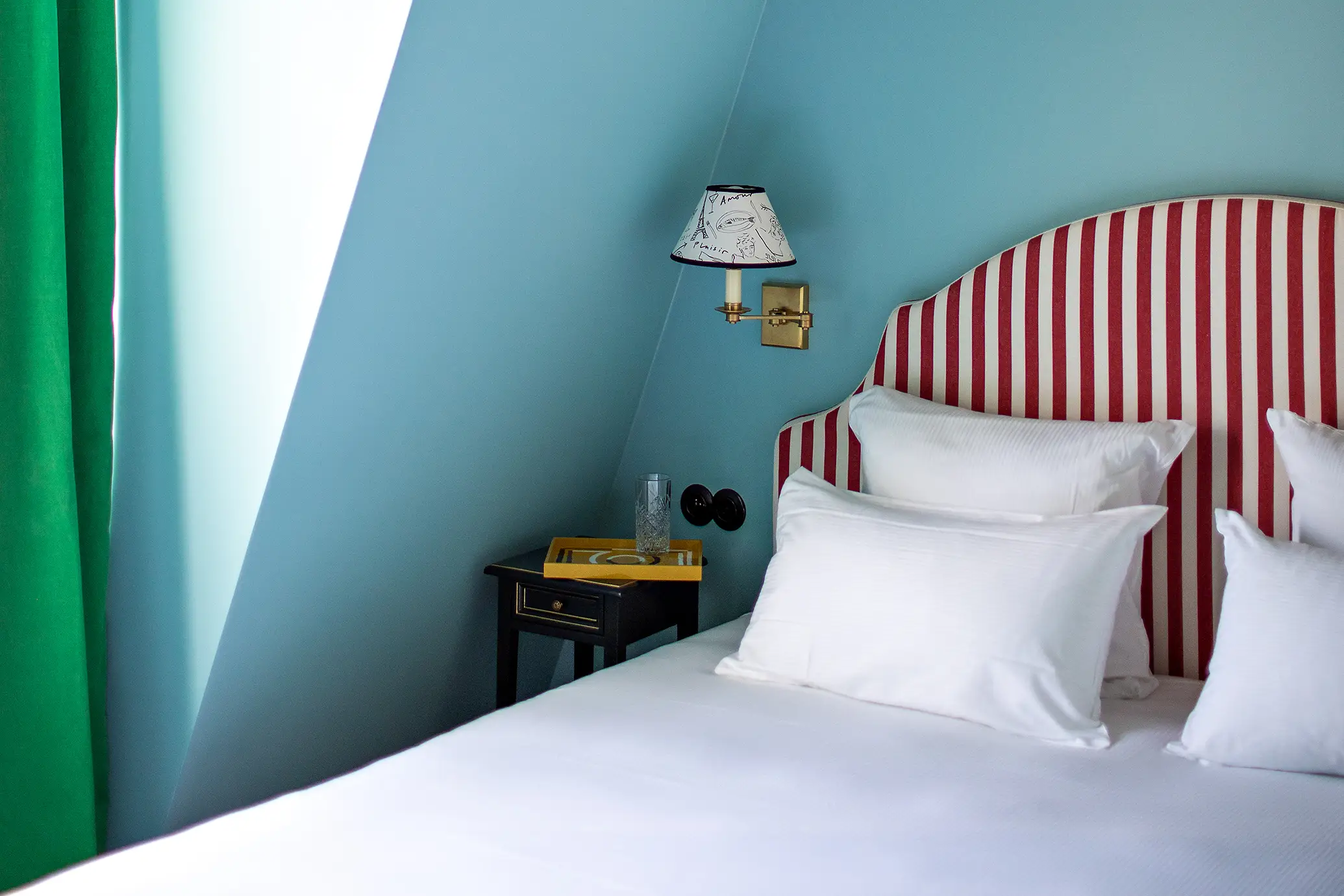 Bed with white pillows tucked under the slope of a blue bedroom, featuring a red-striped headboard, a black nightstand with a yellow top, and an emerald green curtain.