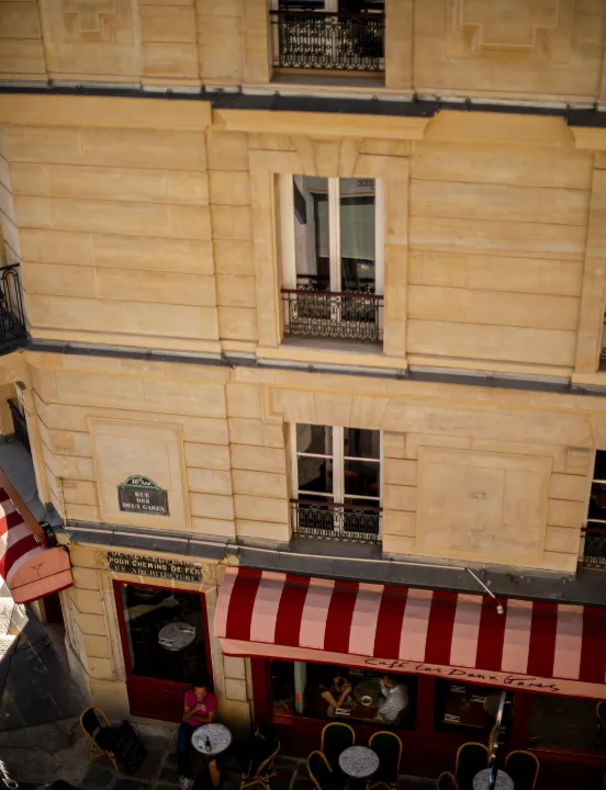Façade en pierre vue d'en haut avec la terrasse animée du Café Les Deux Gares, son store rayé rouge et rose et ses petites tables de bistrot