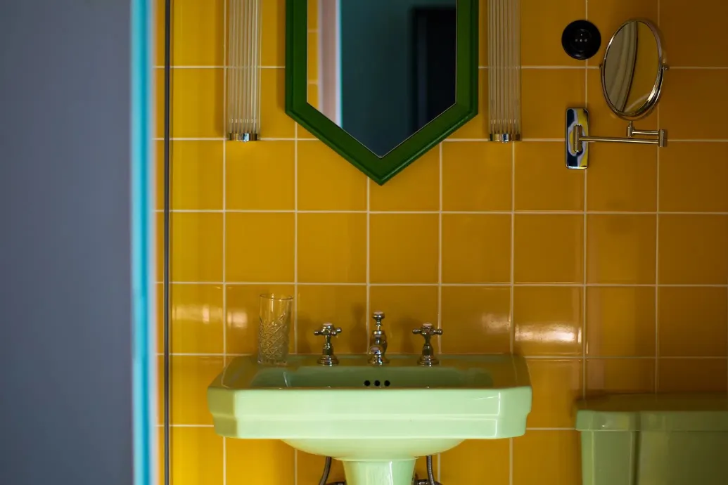 Close-up of an almond green sink with crosshead faucets, topped by a hexagonal green mirror and fluted glass wall sconces on a yellow tiled wall, featuring an articulated magnifying mirror.