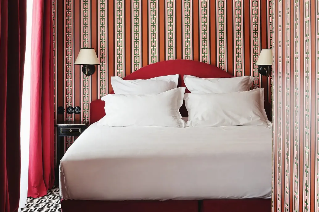 Double bed with white linens nestled in an alcove lined with terracotta stripes and green chains, featuring a red headboard, matching wall sconces, and a crimson velvet curtain