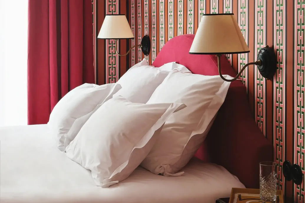 Close-up of white ruffled pillows against a raspberry headboard, beneath two wall lamps with cream shades and in front of a red curtain