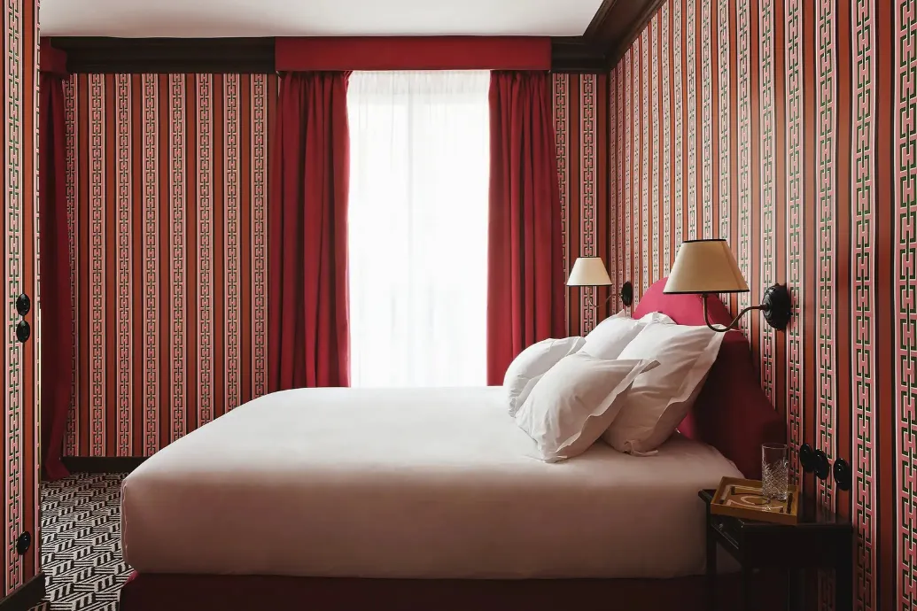Hotel room with red decor, geometric patterned wallpaper, a large bed with white linens, a tufted headboard, wall sconces, and red curtains framing a bright window