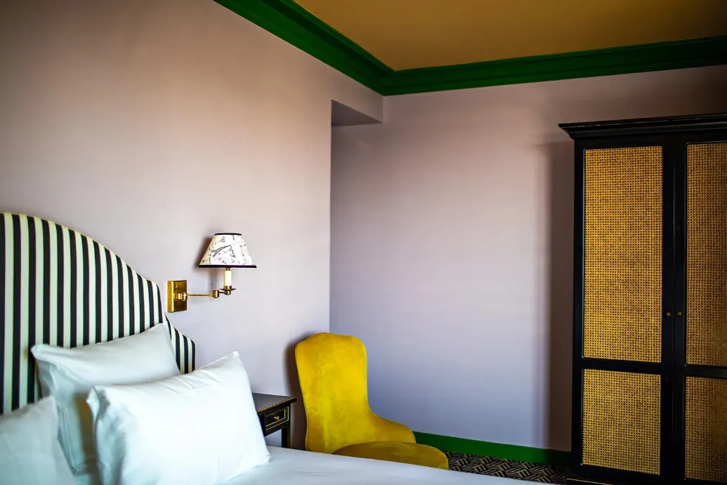 Parma-colored room with an ochre ceiling and green moldings, featuring a black and white striped headboard, a mustard velvet armchair, and a black wardrobe with cane doors