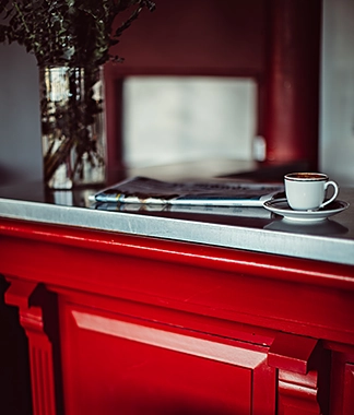 Une petite tasse de café sur une soucoupe, posée sur un comptoir en marbre, accompagnée d'un journal et d'une plante dans un vase en verre, hôtel Les Deux Gares, 4 étoiles, Paris.