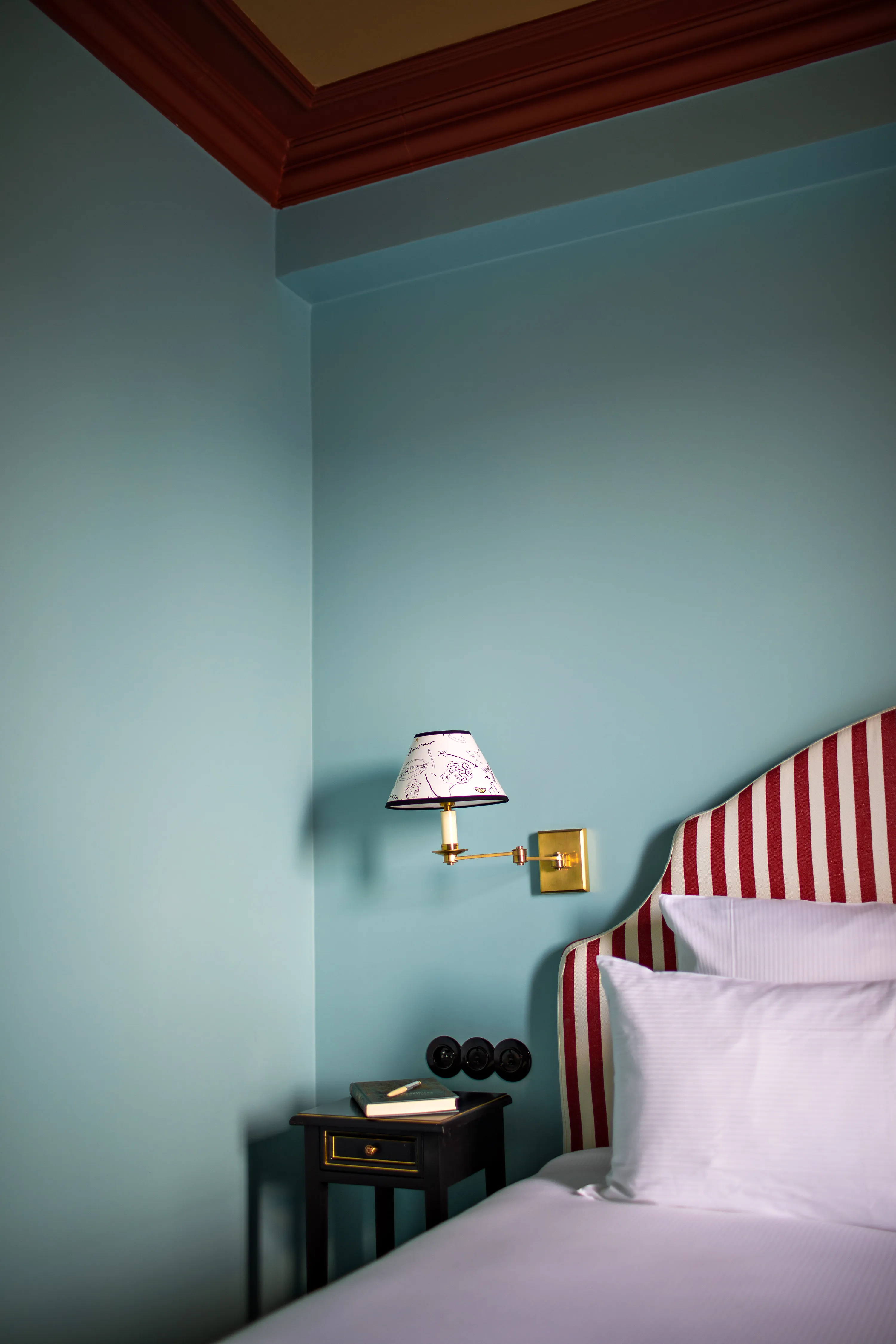Coin d'une chambre avec un mur bleu, une tête de lit rayée rouge et blanche, un lit avec oreillers blancs, une lampe murale dorée avec un abat-jour décoré, et une petite table de chevet noire avec un livre et un crayon dessus.