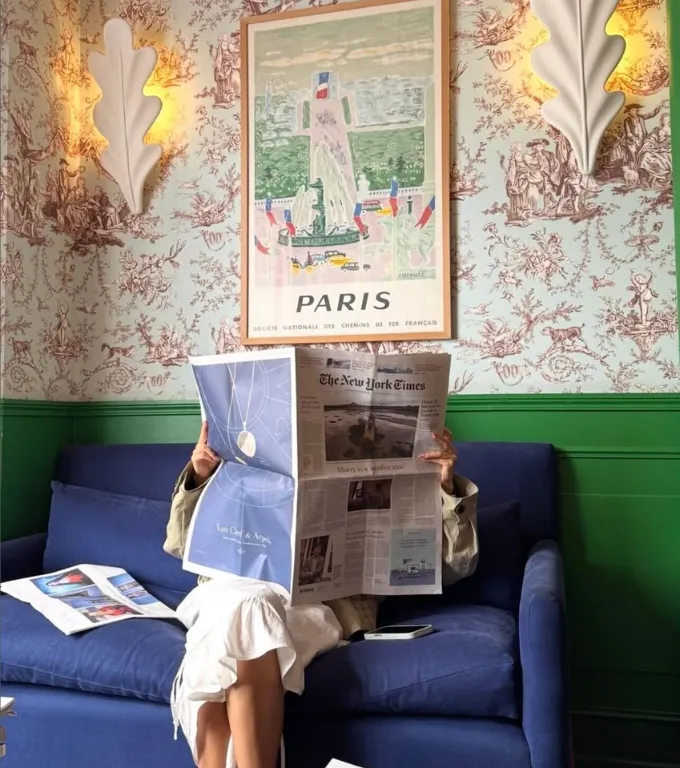 Une personne assise sur un canapé bleu lit le journal The New York Times, cachant son visage. Le mur derrière elle est décoré d'un papier peint avec des motifs rouge foncé et deux appliques murales blanches en forme de feuilles d'acanthe éclairées. Une affiche encadrée au centre montre une illustration colorée de Paris avec des bâtiments et le texte "PARIS" en bas.