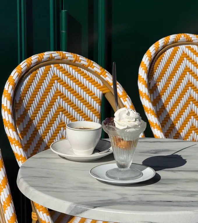 Table ronde en marbre avec une tasse de café blanc sur une soucoupe et un dessert en verre garni de crème fouettée, avec deux chaises tressées en blanc et orange en arrière-plan.