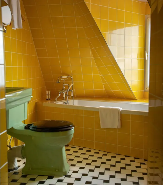 Salle de bain avec carrelage mural jaune vif, baignoire encastrée avec robinetterie rétro, toilette vert clair avec siège noir, et sol en damier noir et blanc.