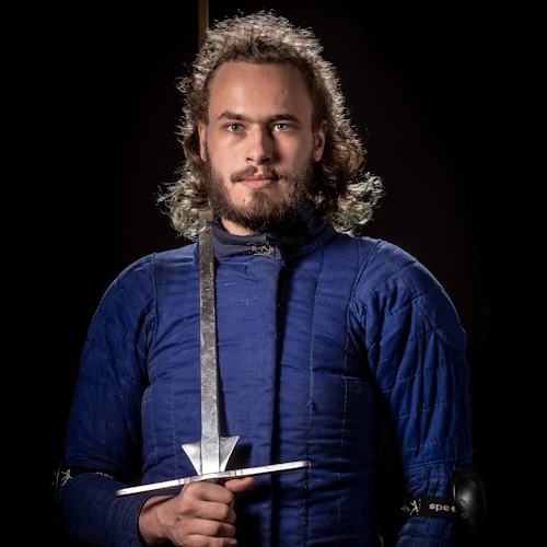 Portretfoto van instructeur Nick Bos. Hij draagt een blauwe jas en draagt een zwaard op zijn schouder.