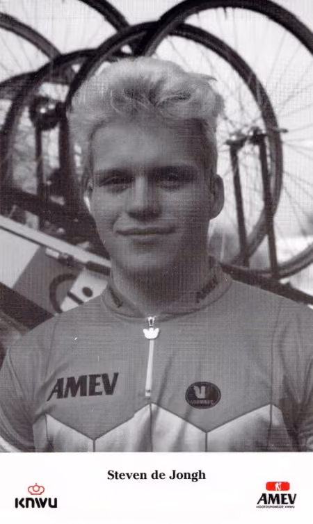 Black and white portrait of a young male cyclist wearing an AMEV jersey, bicycle wheels in background.