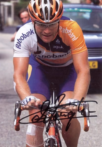 Steven de Jongh in Rabobank jersey and helmet riding a bicycle on a road with a car behind.