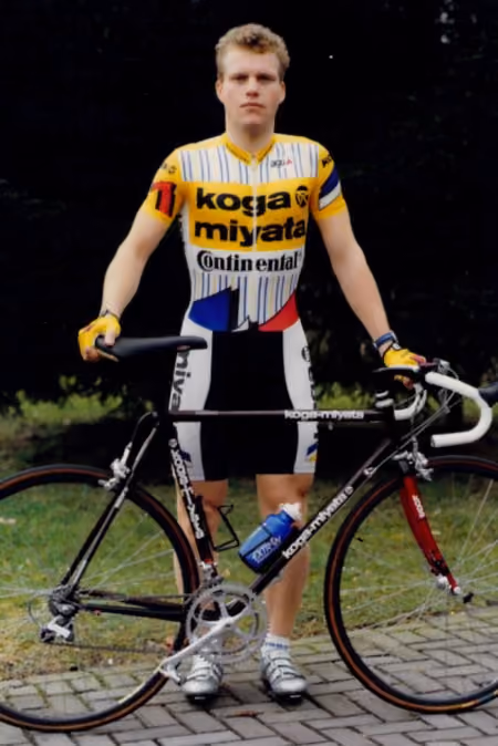Steven de Jongh in yellow Koga Miyata kit standing behind a black road bike on a paved path.