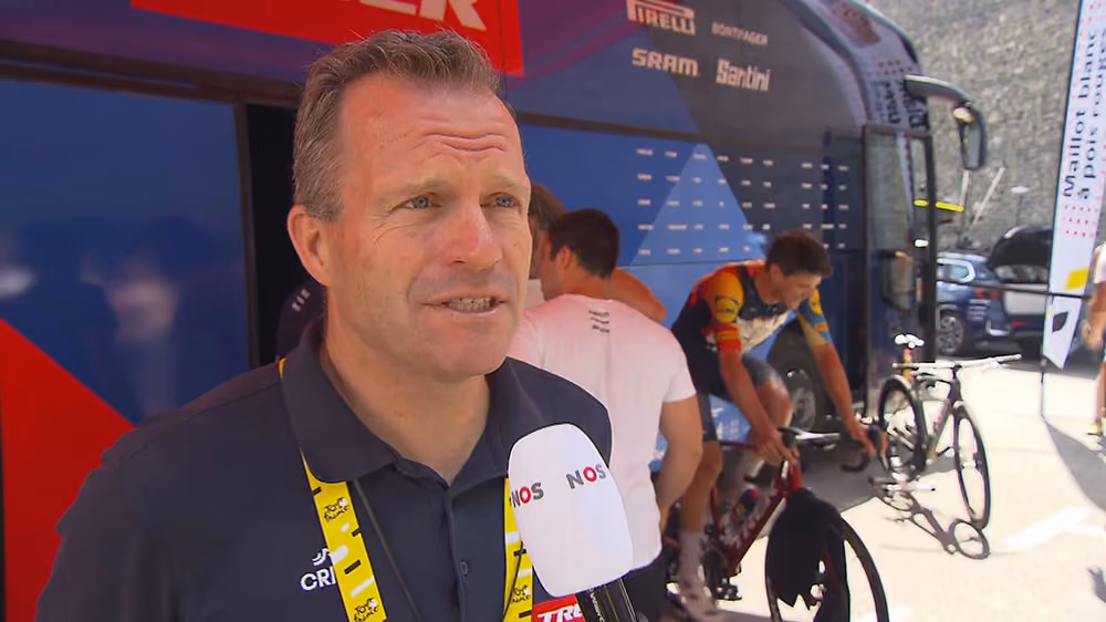 Steven de Jongh wearing a navy shirt and yellow lanyard is interviewed with a microphone near a cycling team bus.