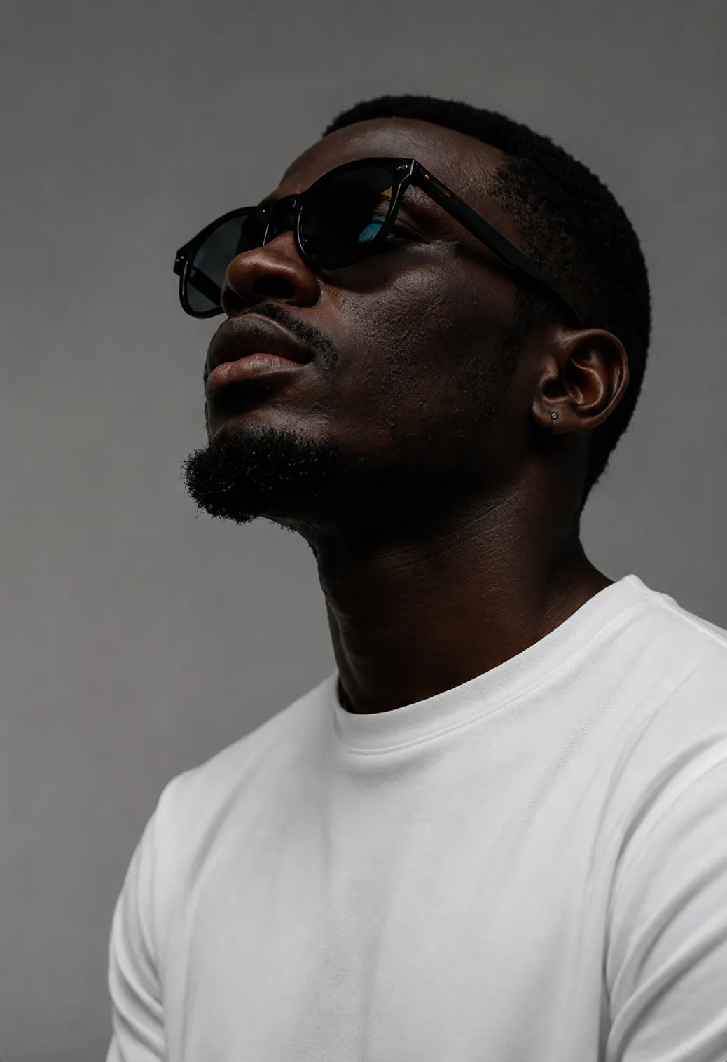 A man with short beard wearing black sunglasses and a white shirt looking upward against gray background.