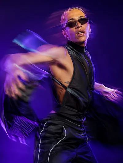 Person in black outfit and sunglasses dancing energetically against a purple background with motion blur.