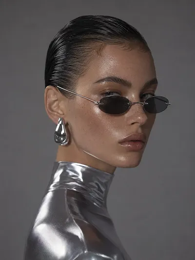 Woman in shiny silver top wears small oval sunglasses and large silver earrings with sleek wet hair.