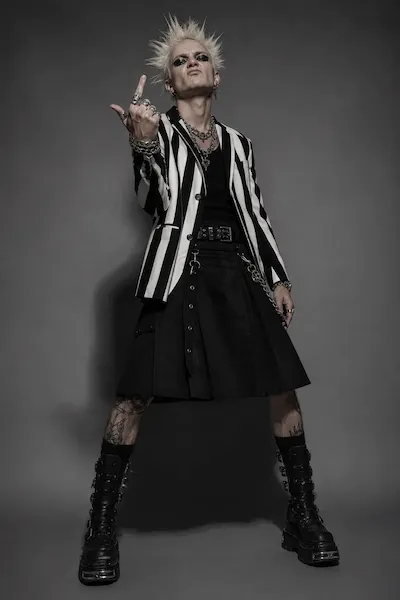 Person with spiky blond hair and heavy makeup wears striped blazer, black kilt, and platform boots.