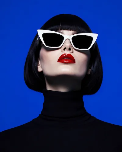 Woman with black bob, white cat-eye sunglasses, red lipstick, and black turtleneck on blue background.
