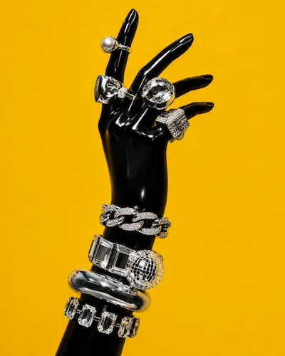 Black mannequin hand with multiple silver rings and bracelets against a yellow background.