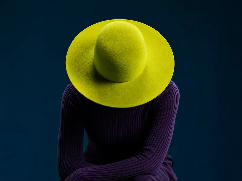 Person with head down wearing a large yellow hat and a dark purple ribbed sweater against a dark background.