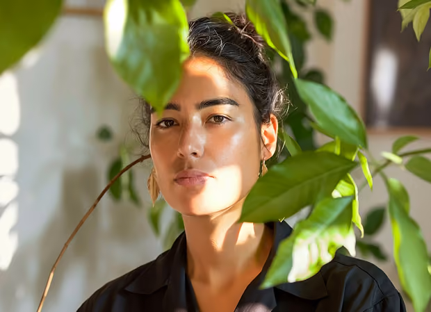 Woman with dark hair and hoop earrings looks at camera with green leaves partially covering her face.