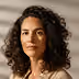 Portrait of a woman with curly hair wearing a beige top against a light background.