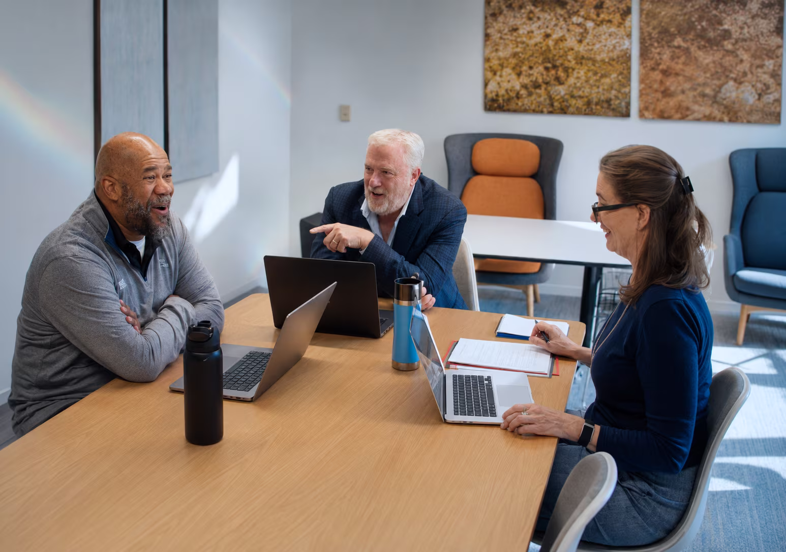 Three colleagues smiling and discussing at a table with laptops and notebooks in a modern office.