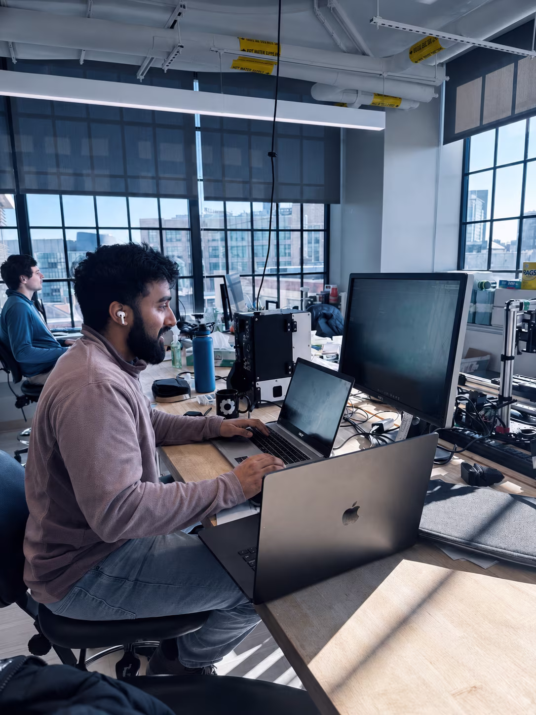 Man wearing earbuds working on multiple laptops and a desktop computer in a bright office.