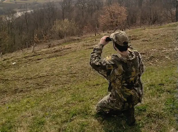 Stefan in camouflage clothing kneeling on grass looking through binoculars toward a wooded valley.