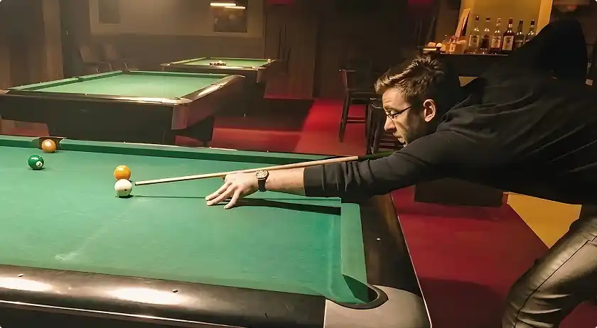 Stefan bending over a pool table lining up a shot with a cue stick in a dimly lit room.