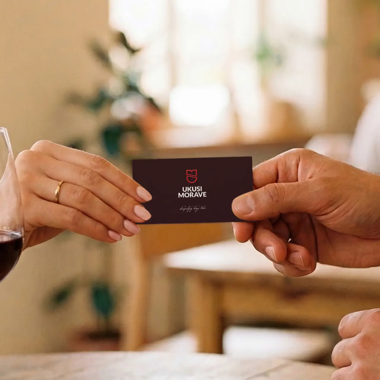 Two hands exchanging a black card reading 'UKUSI MORAVE' indoors with a glass of red wine nearby.