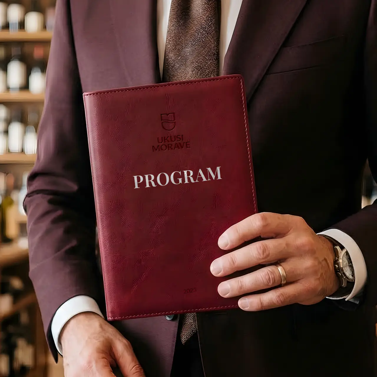 Man in brown suit holding a red leather program book titled 'Ukusi Morave PROGRAM 2023'.