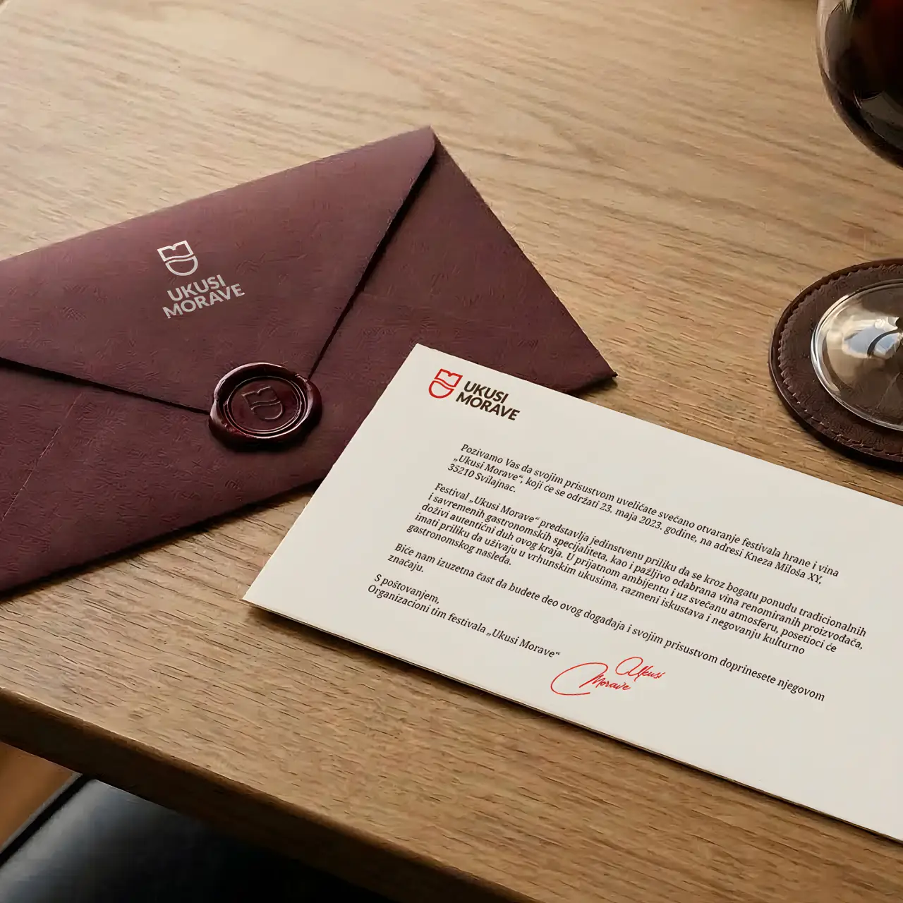 Invitation card and brown envelope with wax seal for Ukusi Morave festival on wooden table.